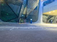 an empty parking lot near a tall building with windows that have lights on it and a car parked in front