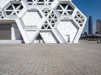 large geometric building with white and silver panels near a plaza with benches under it and cityscape behind