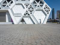 large geometric building with white and silver panels near a plaza with benches under it and cityscape behind