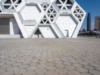 large geometric building with white and silver panels near a plaza with benches under it and cityscape behind