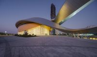 a large building with a curved entrance at night with an interesting design and glass tower in the distance