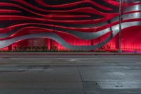 a city street has a large wavy building with bright red lights above it and a sidewalk at night