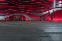 a city street has a large wavy building with bright red lights above it and a sidewalk at night