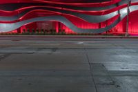 a city street has a large wavy building with bright red lights above it and a sidewalk at night