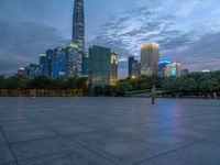 a paved patio surrounded by tall buildings at dusk time with a bench to sit on