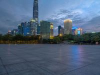 a paved patio surrounded by tall buildings at dusk time with a bench to sit on