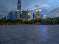 a paved patio surrounded by tall buildings at dusk time with a bench to sit on