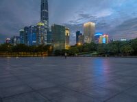 a paved patio surrounded by tall buildings at dusk time with a bench to sit on