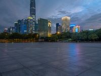a paved patio surrounded by tall buildings at dusk time with a bench to sit on