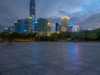 a paved patio surrounded by tall buildings at dusk time with a bench to sit on