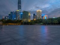 a paved patio surrounded by tall buildings at dusk time with a bench to sit on