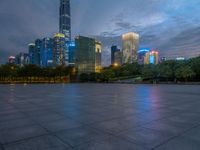 a paved patio surrounded by tall buildings at dusk time with a bench to sit on