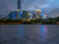 a paved patio surrounded by tall buildings at dusk time with a bench to sit on