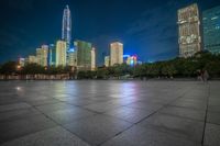 a city skyline with high rise buildings and a person walking around on a paved ground at night