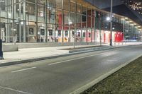 an empty street at night in front of a tall glass building with a bus stop sign