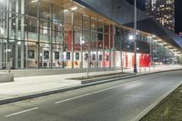 an empty street at night in front of a tall glass building with a bus stop sign