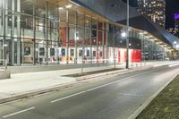 an empty street at night in front of a tall glass building with a bus stop sign