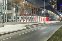 an empty street at night in front of a tall glass building with a bus stop sign