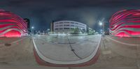 360 lens photograph of a parking lot at night time and buildings near by by and there is a bright red light on the roof