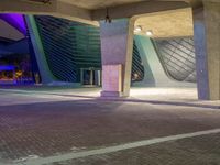 an empty parking area has the building lit up purple and blue at night with a few people walking