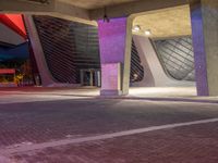 an empty parking area has the building lit up purple and blue at night with a few people walking