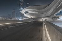 the view of a large, curved building over an empty road at night with urban buildings in the background