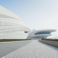 a walkway leading to a modern building with curved shapes on it and white walls, and blue windows in the sky