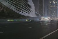 a large building sitting near a highway with skyscrapers in the background on a dark night