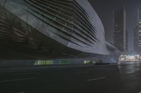 a large building sitting near a highway with skyscrapers in the background on a dark night