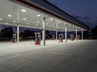 several gas pumps sit on concrete at a station with lights above them on an empty street