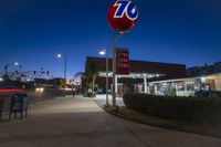 street signs in front of a gas station on a city street at night with people standing under a traffic light