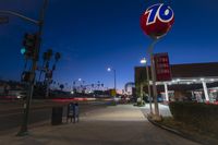 street signs in front of a gas station on a city street at night with people standing under a traffic light