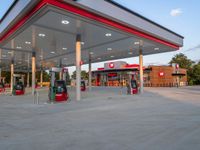 gas station at the edge of a street next to a large white building with red trimming