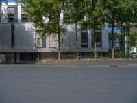 a large building with an entrance on a city street lined by tall green trees, and a small bench near it