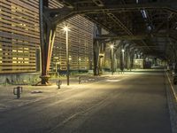 an empty road leading to a large train station at night time by a row of parking cars and a wooden wall in the background