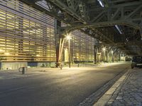 an empty road leading to a large train station at night time by a row of parking cars and a wooden wall in the background