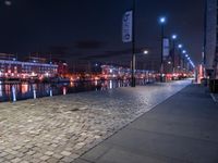 a long street lined with brick walkway next to a river at night time with street lights and city lights