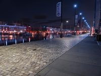 a long street lined with brick walkway next to a river at night time with street lights and city lights