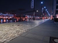 a long street lined with brick walkway next to a river at night time with street lights and city lights