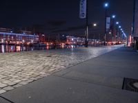 a long street lined with brick walkway next to a river at night time with street lights and city lights