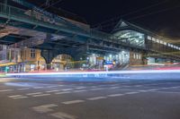 an open road with cars going by it at night, including a pedestrian overpass and two train bridges at the top of it