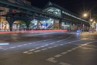 an open road with cars going by it at night, including a pedestrian overpass and two train bridges at the top of it