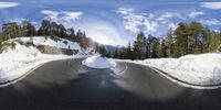 a snow covered mountain side next to a winding road with trees in the background and a big sky with some clouds