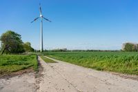 a single windmill standing on top of a green field next to a road, near a small dirt road