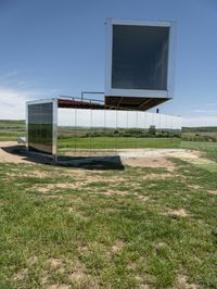 a mirrored structure sits on a field near some grass and a lake in the background