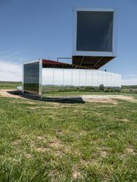 a mirrored structure sits on a field near some grass and a lake in the background