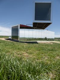 a mirrored structure sits on a field near some grass and a lake in the background