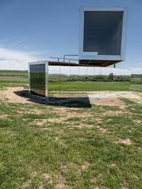 a mirrored structure sits on a field near some grass and a lake in the background