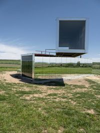 a mirrored structure sits on a field near some grass and a lake in the background