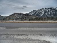 snow covering the ground with mountains in the background on a cloudy day, near an empty road and fence